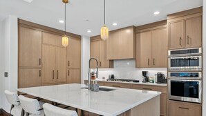 Uma cozinha moderna com armários de madeira, uma ilha central, eletrodomésticos de aço inoxidável e iluminação pendente.