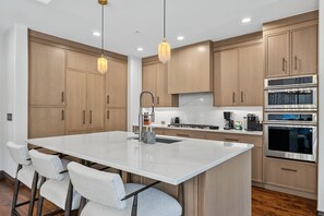A modern kitchen with wooden cabinets, a central island, stainless steel appliances, and pendant lighting. - Contemporary Condo by InvitedHome | Ski-In/Out, Base of Village, Balcony, Views (Avon)