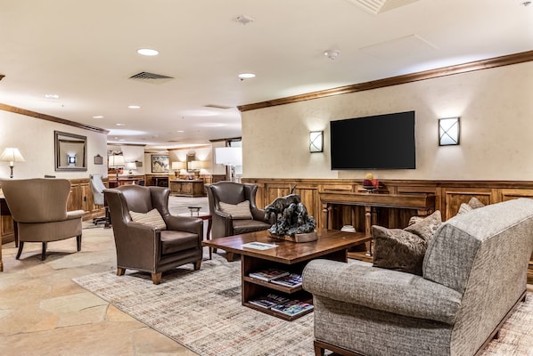 A spacious living room with leather armchairs, a coffee table with magazines, a flat-screen TV mounted on the wall, and a wooden accent wall.