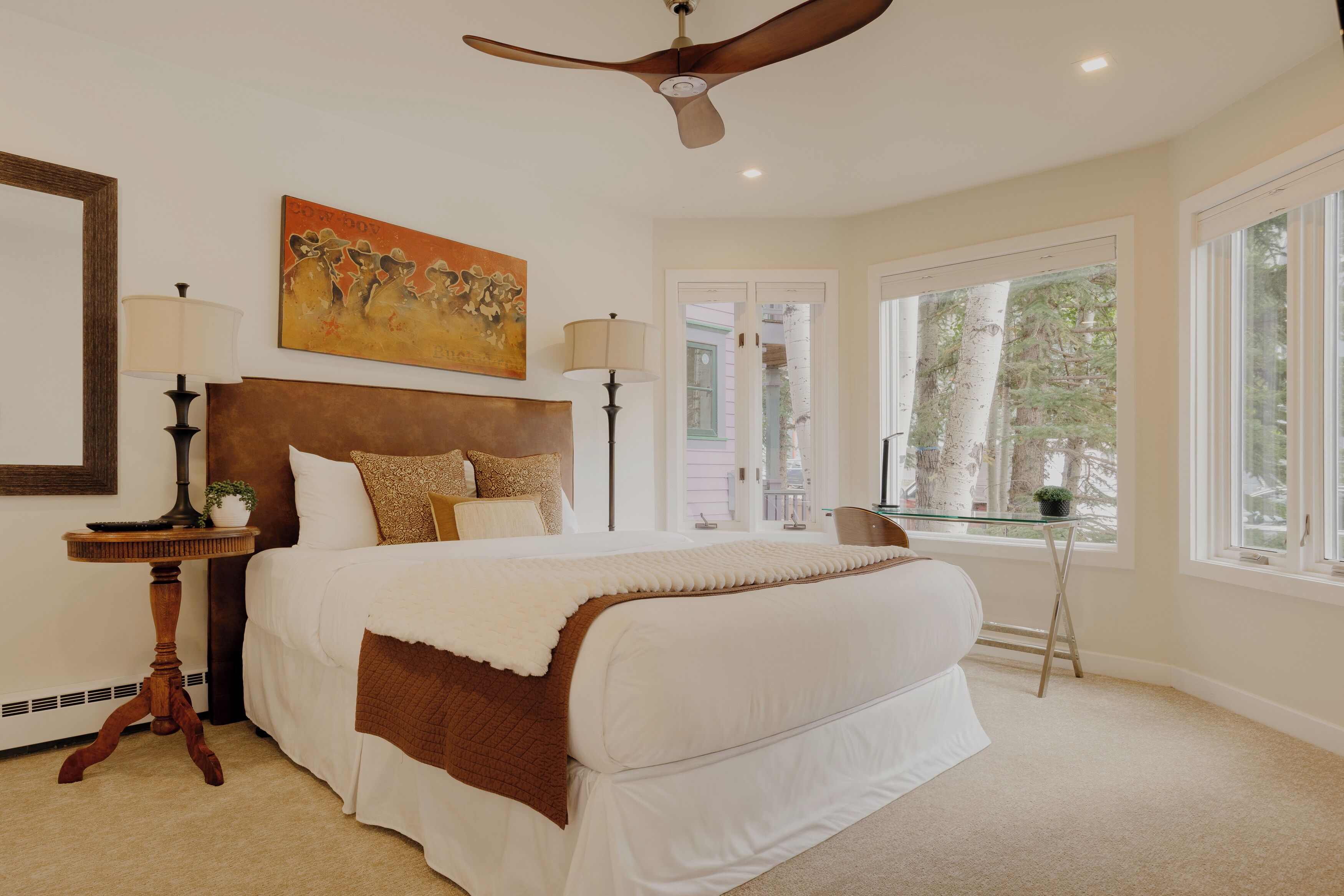 A bedroom with a large bed, a ceiling fan, a painting, and a window with a view of trees.