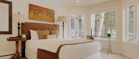 A bedroom with a large bed, a ceiling fan, a painting, and a window with a view of trees.