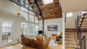 Un salon spacieux avec un plafond en bois, un canapé en cuir marron, un escalier en bois et de grandes fenêtres.