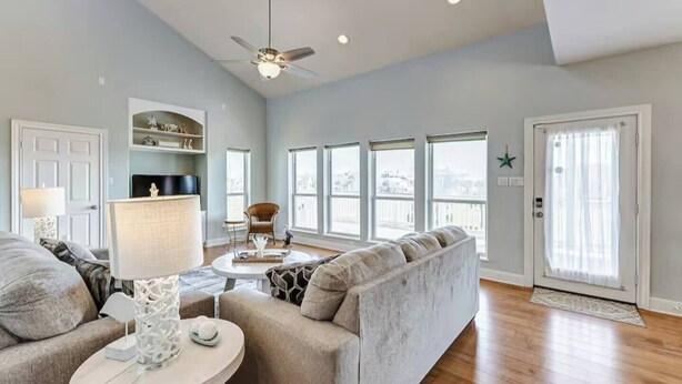 A spacious living room with a high ceiling, a ceiling fan, a fireplace, a sofa set, a coffee table, and large windows offering a view.