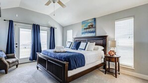 A bedroom with a large bed, a ceiling fan, a painting of a pier, and a view of the outdoors through the window.