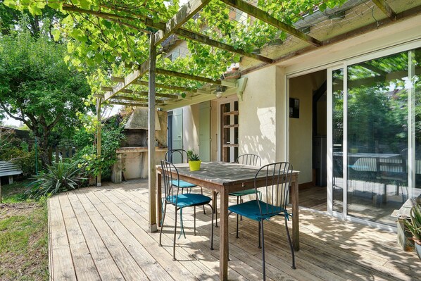 A wooden deck with a table and chairs, a pergola with climbing plants, and a sliding glass door leading to a covered patio area.