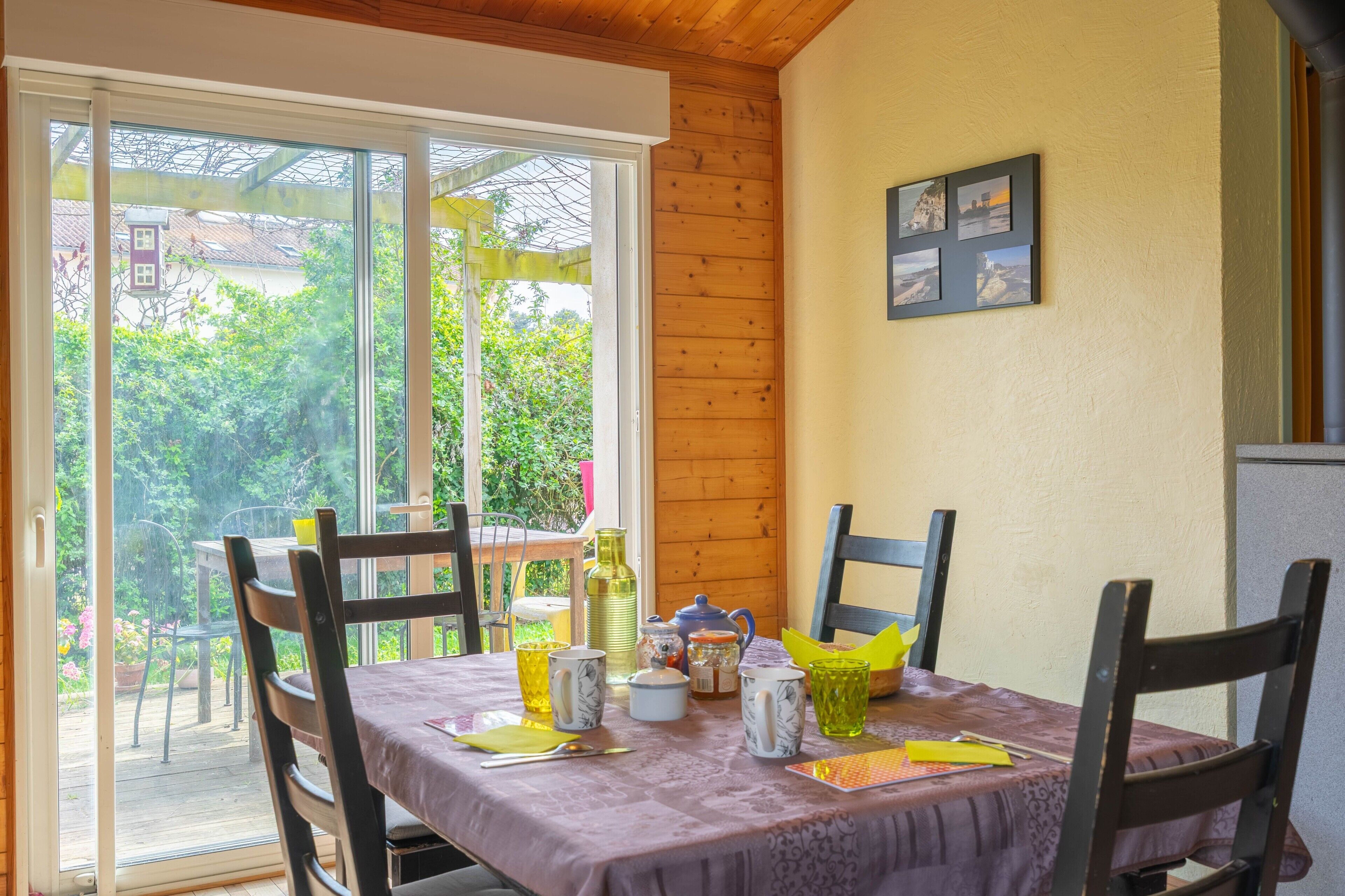 A dining area with a table set for two, chairs, a window with a view of greenery, and a wooden wall.