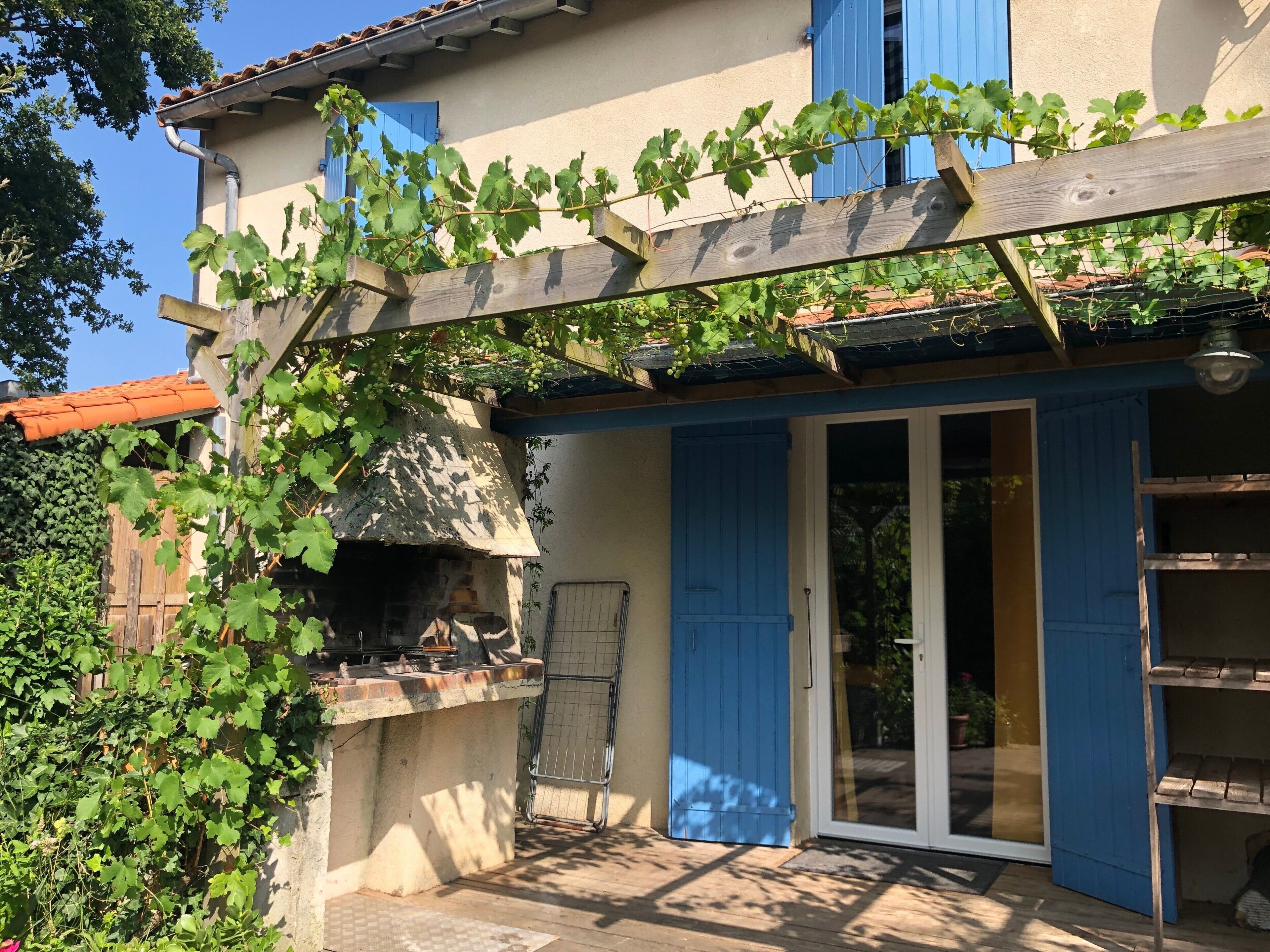 Un bâtiment doté d’une cheminée en pierre, d’une pergola en bois recouverte de plantes grimpantes et d’une porte bleue.