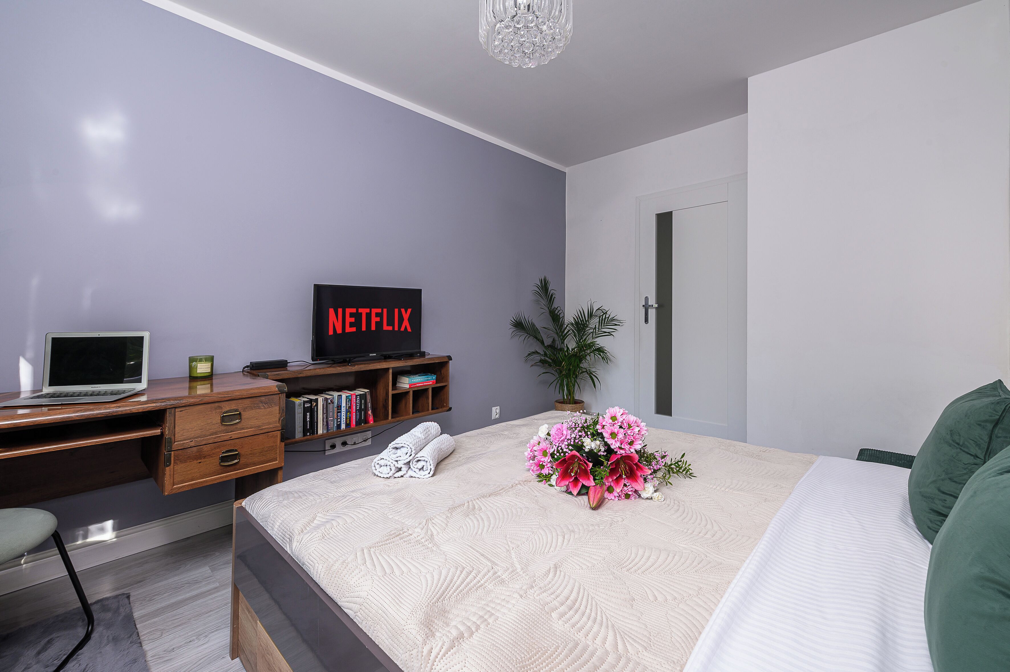 Quarto com cama, escrivaninha com laptop, TV exibindo 'NETFLIX', planta em vaso e um arranjo de flores.