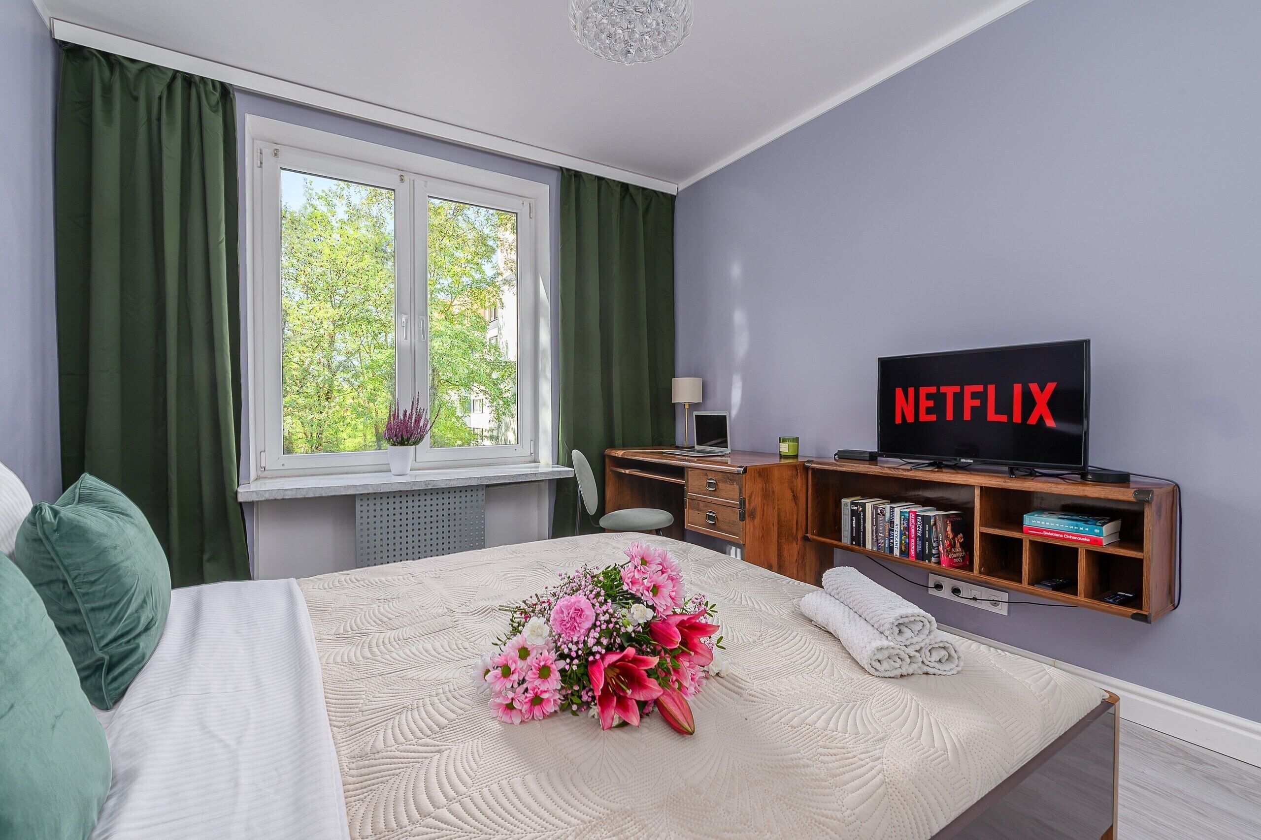 A hotel room with a bed, a desk, a television displaying 'NETFLIX', and a window with curtains.