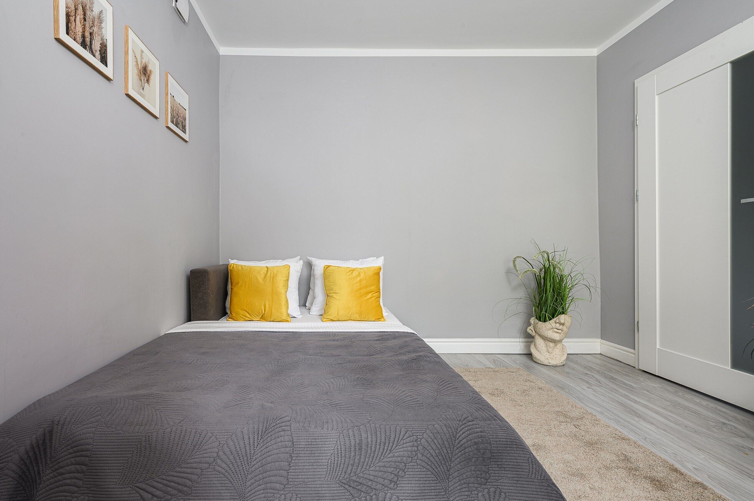 A bedroom with a large bed, two yellow pillows, a grey bedspread, and a plant in a vase.