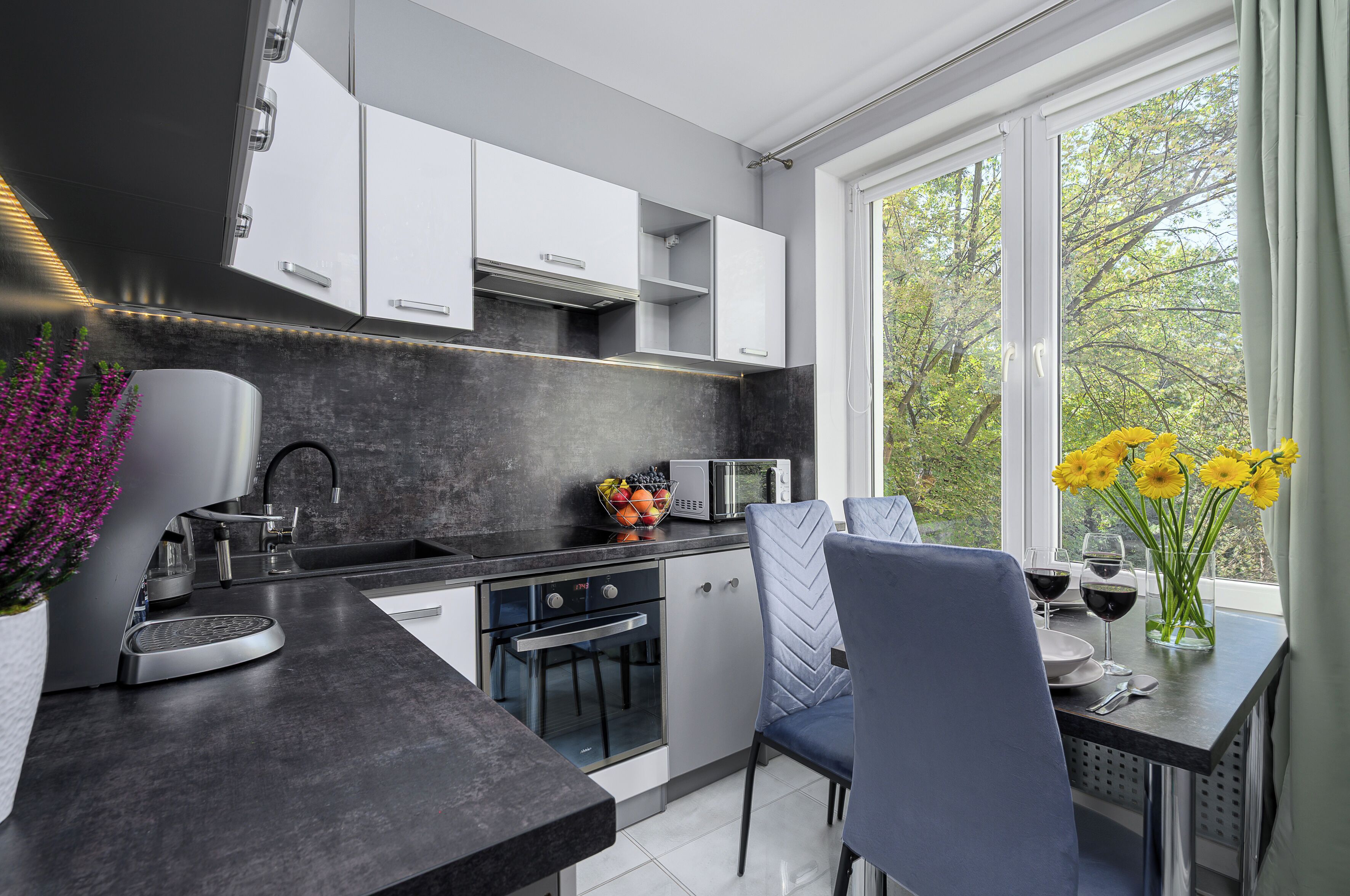 Cocina moderna con horno empotrado, microondas y fregadero. Hay un comedor con mesa y sillas, y una ventana con vistas a árboles.