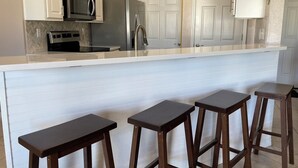 A kitchen with white cabinets, stainless steel appliances, a white countertop, and three wooden stools. - Waterfront Playground in Paradise - more than a condo rental, a destination! (Destin)