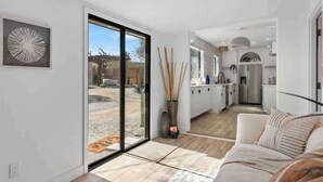 Un intérieur moderne avec une porte-fenêtre coulissante donnant sur un patio, une cuisine avec des meubles blancs et un espace de vie doté d’un canapé.