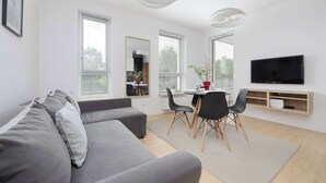 A modern living room with a grey sofa, dining table with chairs, a flat-screen TV, and large windows.
