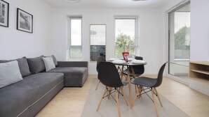 A modern living room with a grey sectional sofa, a dining table with four chairs, and a large window with blinds.