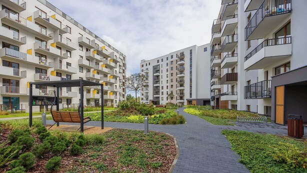 A modern residential building with balconies, a paved walkway, and a small garden area with a bench.