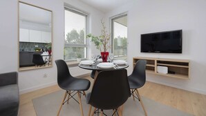 A modern living room with a dining area, a flat-screen TV mounted on the wall, and a kitchen visible through a large mirror.