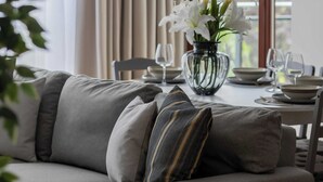 A modern living room with a gray sofa, a dining table set with plates and glasses, and a vase of white flowers.
