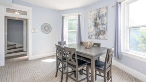 Un coin repas avec une table et des chaises, une fenêtre avec des rideaux, une horloge murale et une porte donnant sur un escalier.