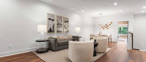 A modern living room with a grey sofa, round ottoman, and a coffee table. There is a rug on the floor, a lamp, and a painting on the wall.