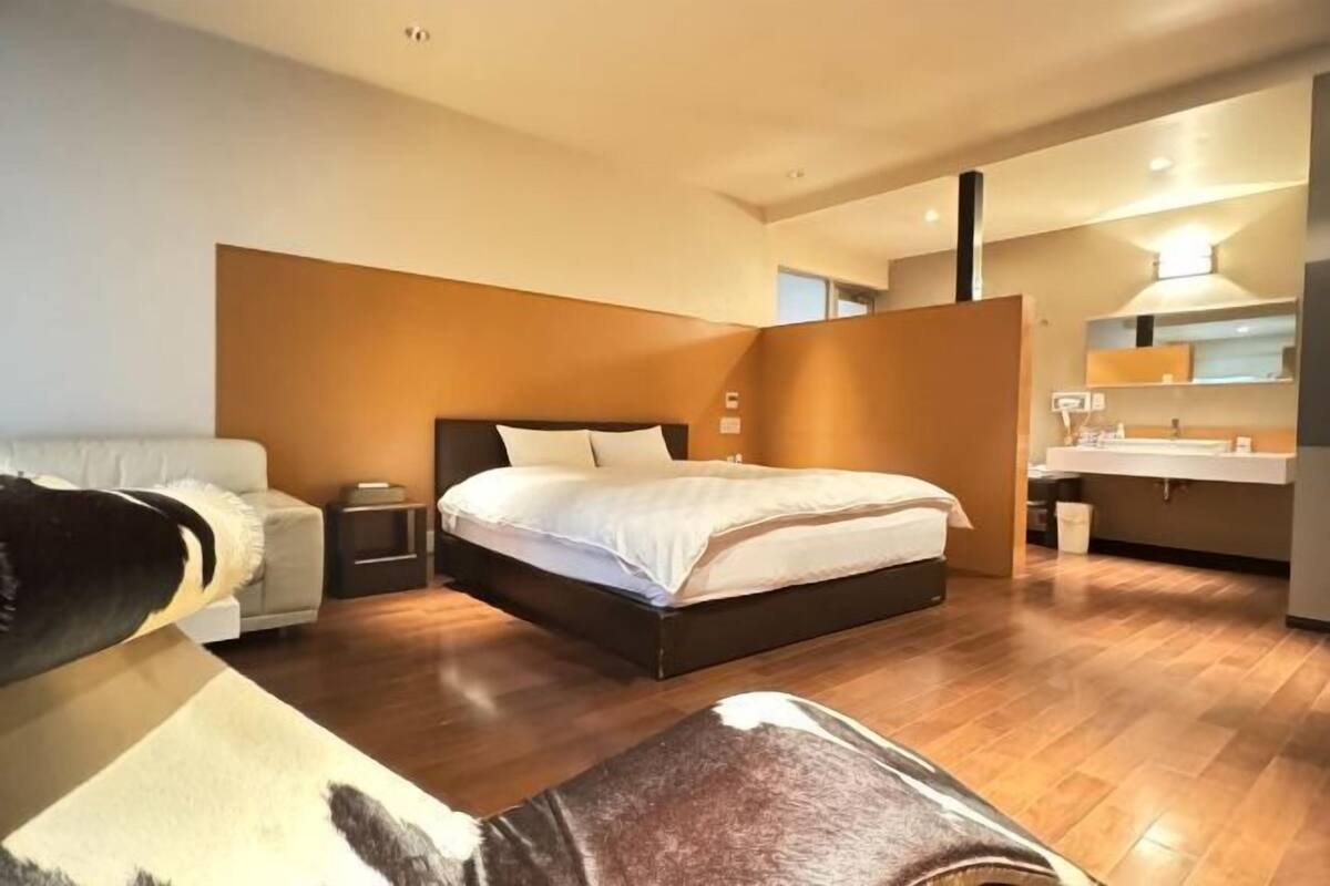 A modern bedroom with a bed, a sofa, a bedside table, and a bathroom with a sink and mirror.