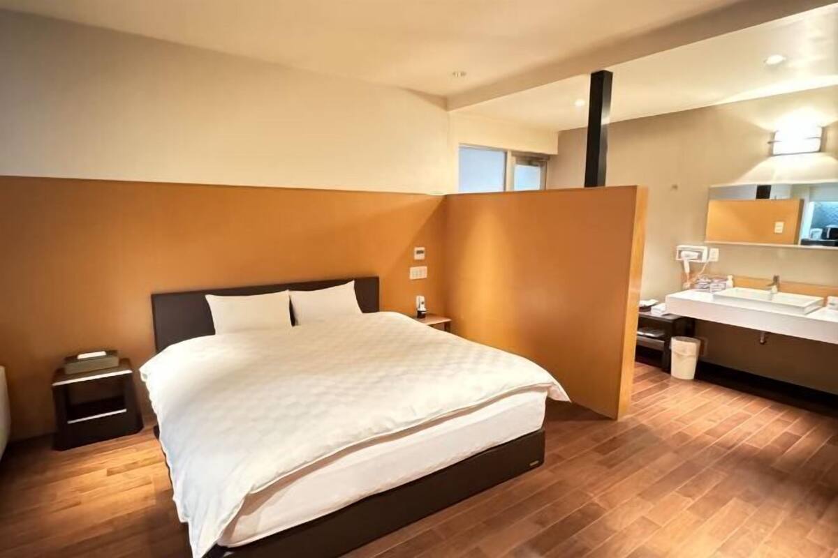A modern hotel room with a large bed, a bedside table, a nightstand, a bathroom with a sink and mirror, and a wooden floor.