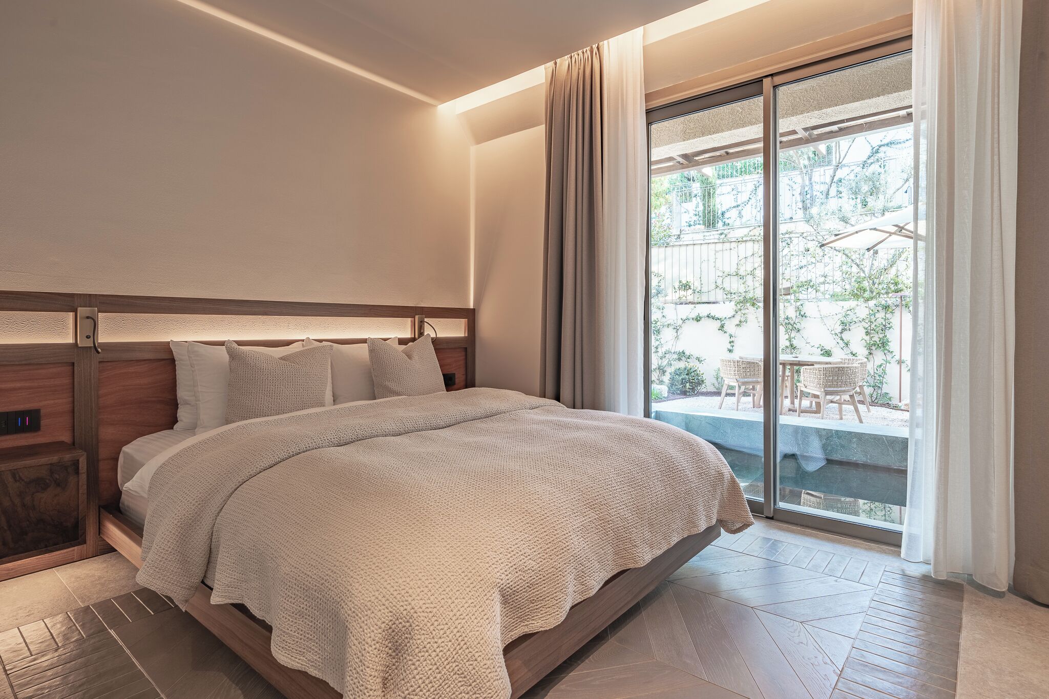 A modern hotel room with a large bed, a wooden headboard, a nightstand, and a sliding glass door leading to an outdoor area.