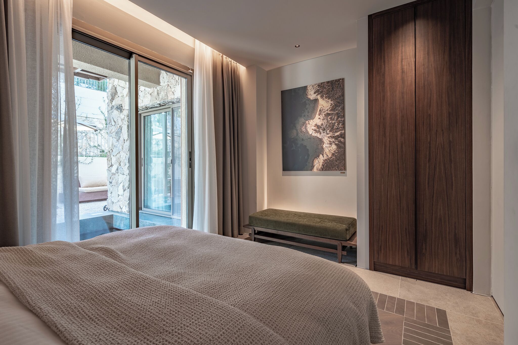 A bedroom with a large bed, a bench, a wooden wardrobe, a window with curtains, and a wall-mounted artwork.