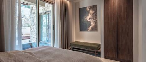 A bedroom with a large bed, a bench, a wooden wardrobe, a window with curtains, and a wall-mounted artwork.
