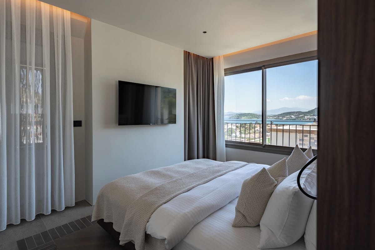 Une chambre d’hôtel avec un lit, un téléviseur fixé au mur, une grande fenêtre donnant sur la mer et des rideaux.