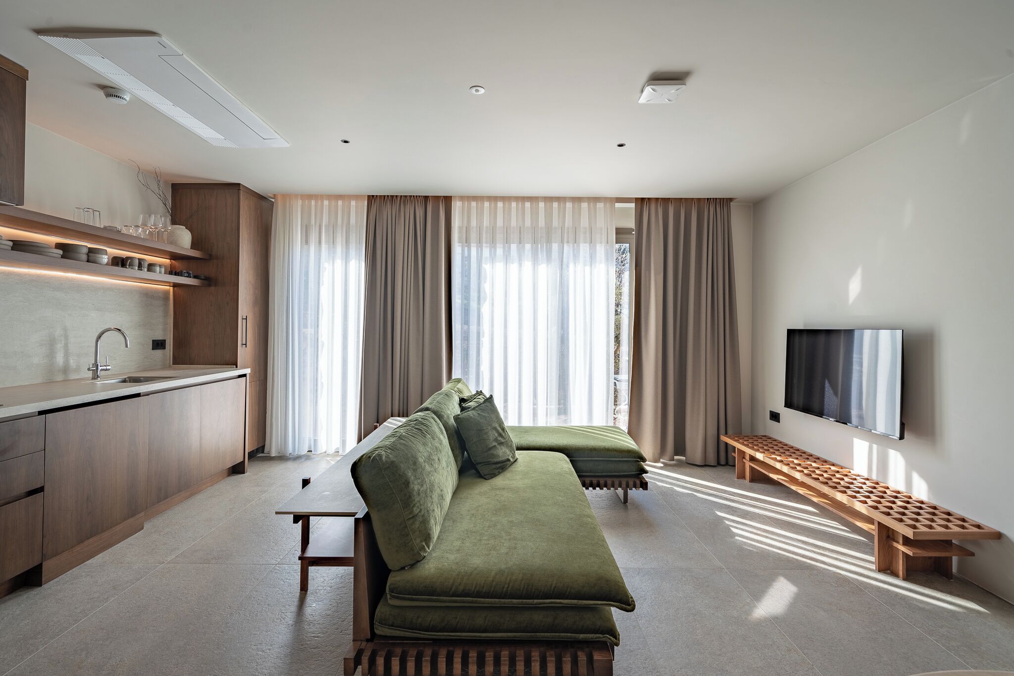 Una sala de estar moderna con un sofá, televisión, zona de cocina y ventanas amplias.