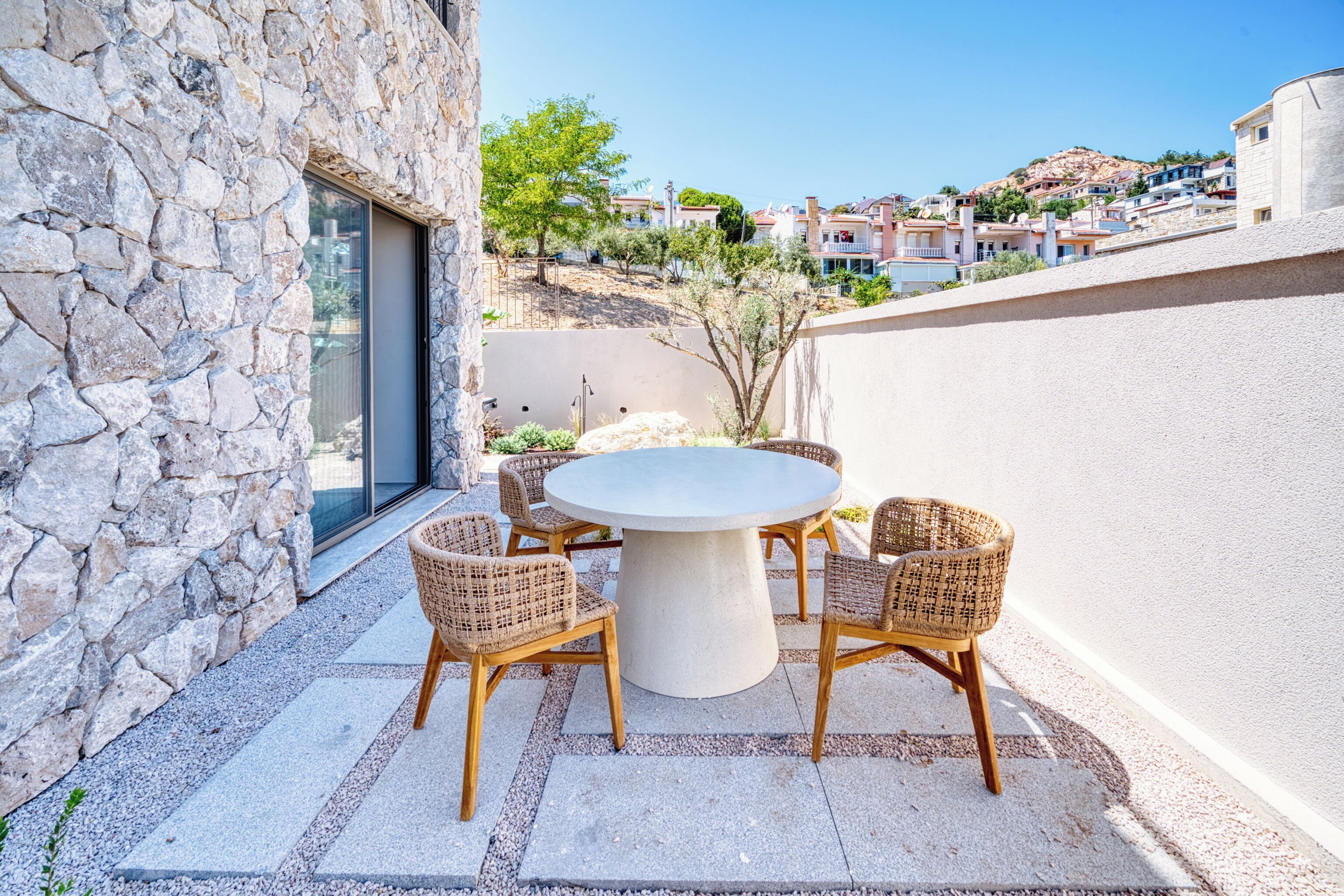 A stone building with a round table and wicker chairs, a glass door, and a view of a hillside with buildings.