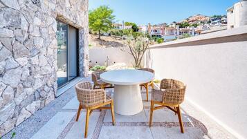 A stone building with a round table and wicker chairs, a glass door, and a view of a hillside with buildings.