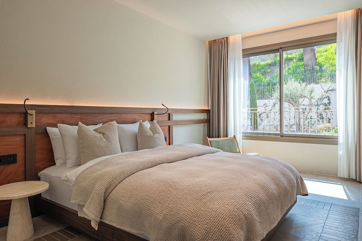 Une chambre d’hôtel avec un grand lit, une table de chevet, une fenêtre avec des rideaux et une vue sur la verdure.