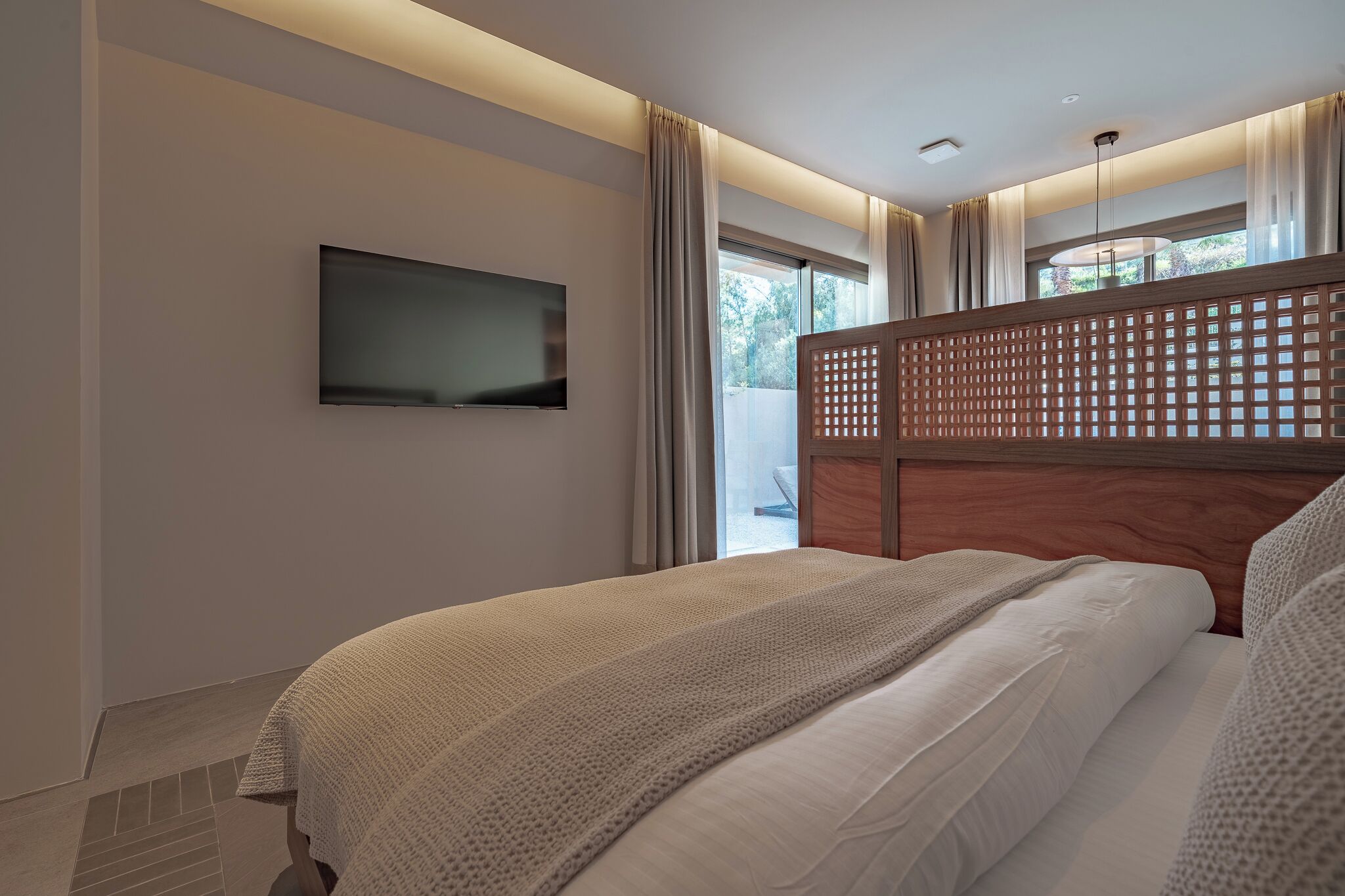 A modern hotel room with a bed, a flat-screen TV mounted on the wall, and a large window with curtains.