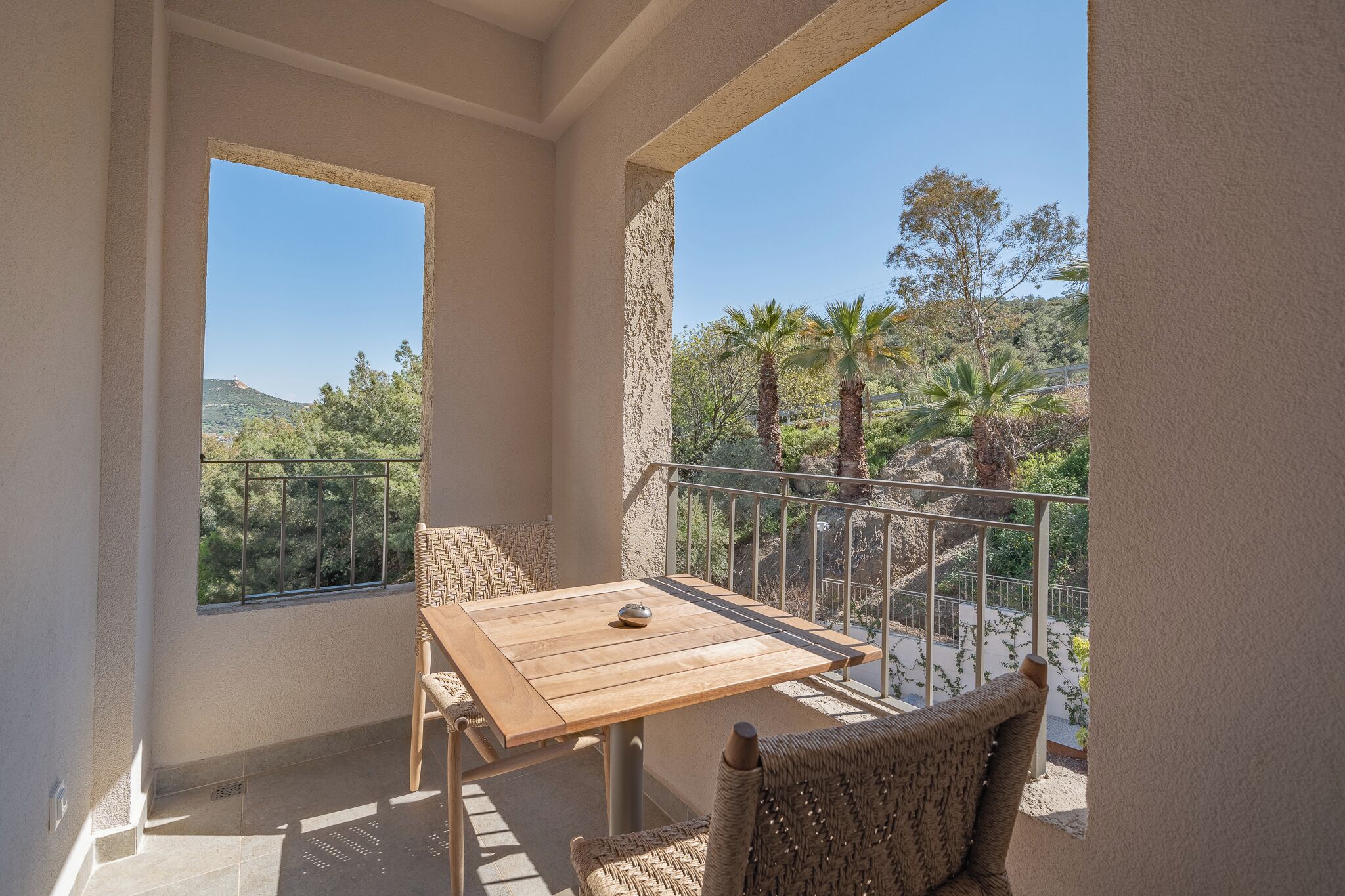 A balcony with a wooden table and wicker chairs, overlooking a mountainous landscape with trees and a clear sky.