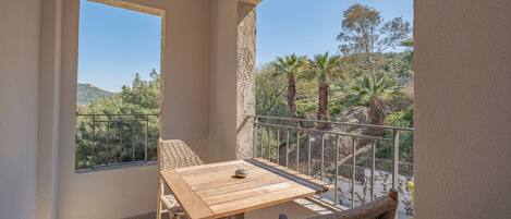A balcony with a wooden table and wicker chairs, overlooking a mountainous landscape with trees and a clear sky.