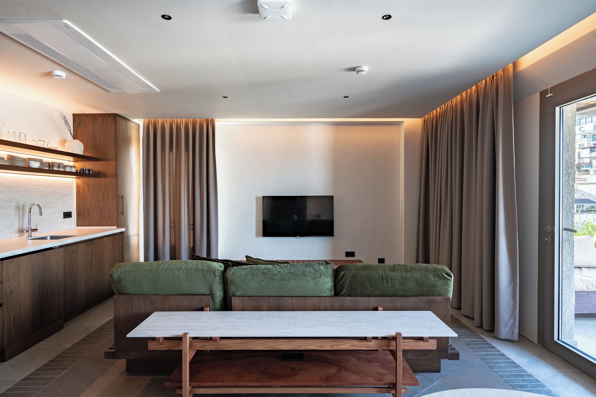 A modern living area with a sofa, a coffee table, a flat-screen TV, and a kitchen area with a sink and cabinets.