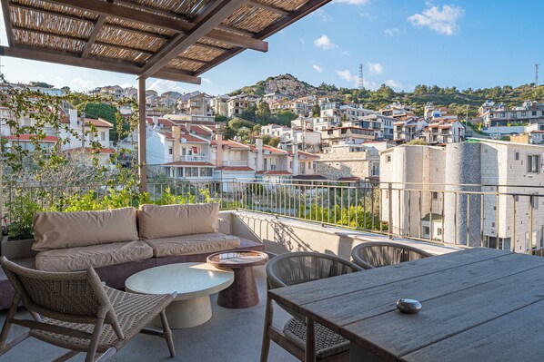 A balcony with a sofa, chairs, and a table, overlooking a hillside town. - Käsiala Apart Hotel Yeni Foça (Foça)