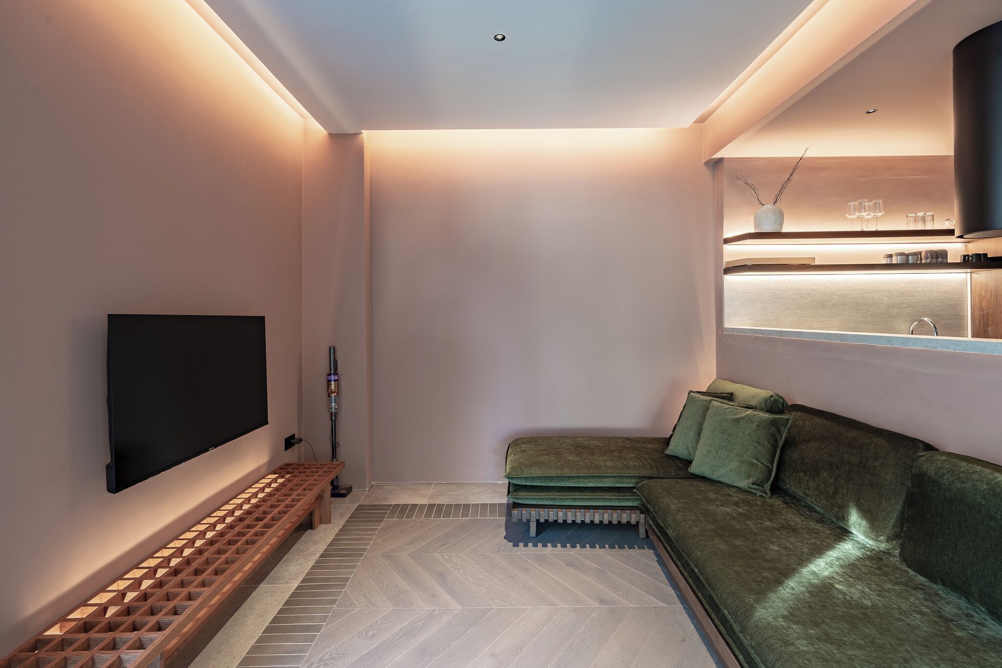A modern living room with a flat-screen TV, a wooden bench, a green sofa, and a kitchen area with a window above.