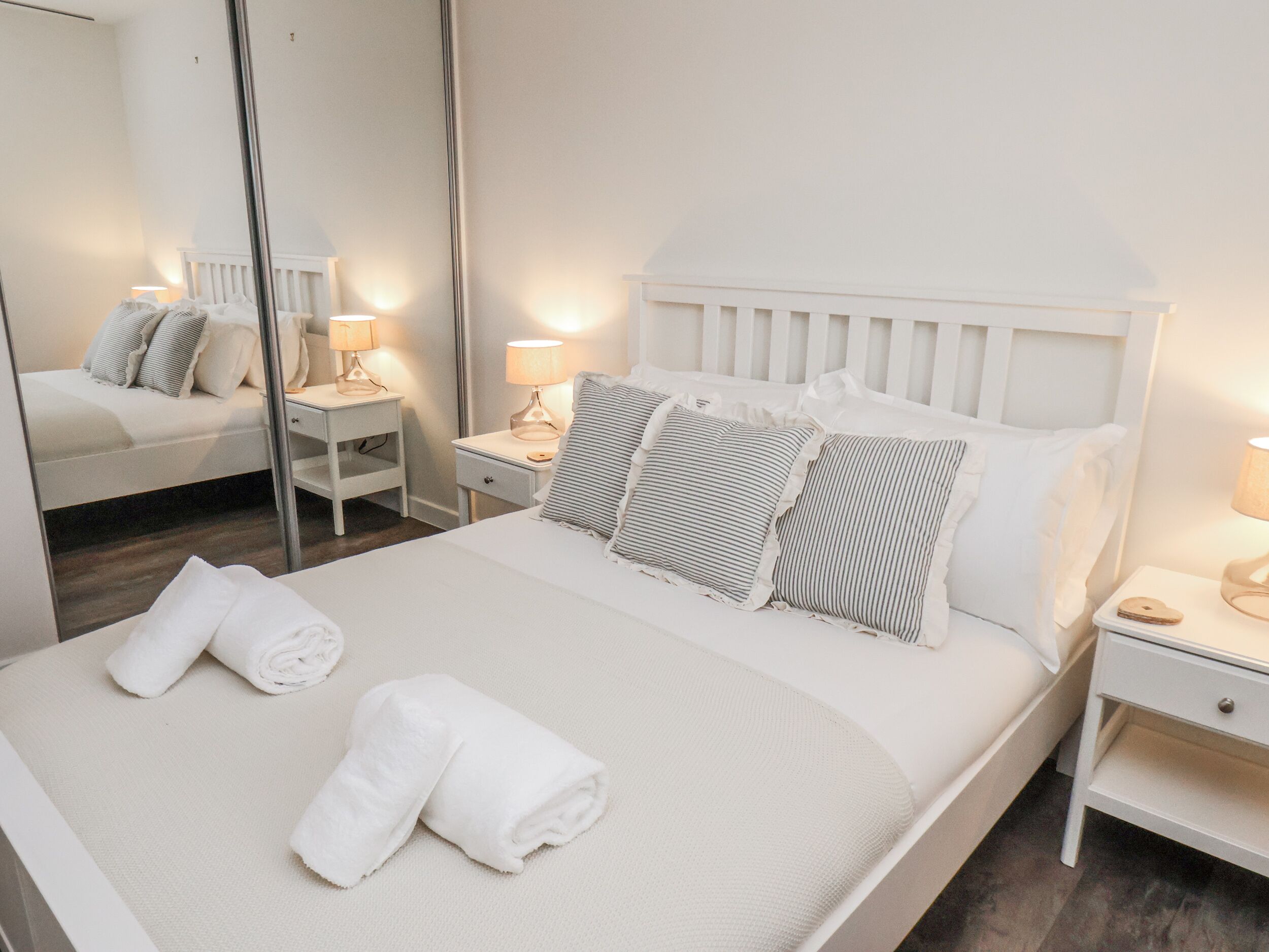 A neatly made bed with white linens and striped pillows, two bedside tables with lamps, and a large mirror on the wall.