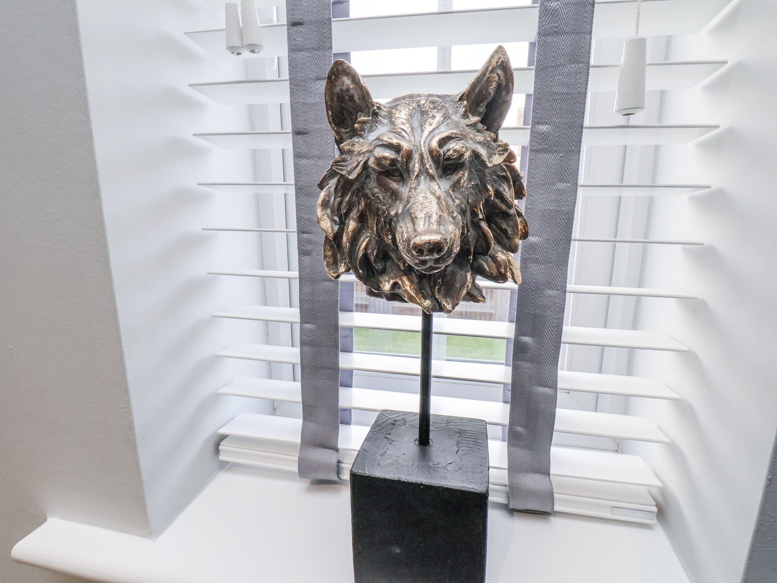 A sculpture of a wolf's head mounted on a black pedestal in front of a window with white blinds.