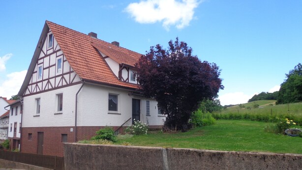 A traditional half-timbered house with a sloped roof, a stone wall in front, and a grassy area with a tree. - Vacation cottage Van Wijk (Alheim)