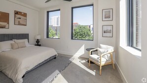 Ein modernes Schlafzimmer mit einem großen Bett, einem Deckenventilator, zwei Fenstern mit Blick auf die Stadt und einem kleinen Nachttisch.