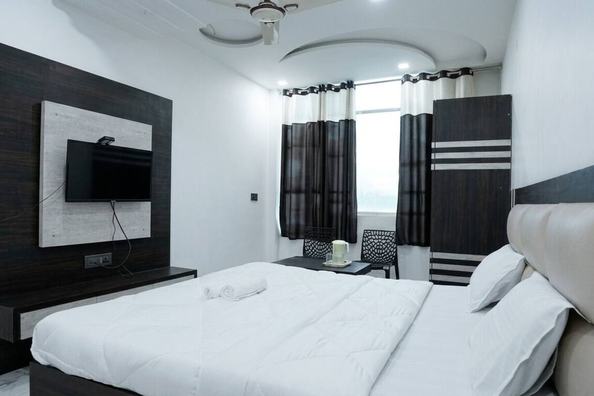 A modern hotel room with a large bed, a flat-screen TV mounted on the wall, a desk with a chair, and a window with curtains.