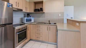 Una cocina compacta con refrigerador de acero inoxidable, microondas y horno empotrado. Hay un lavamanos con grifo, y los armarios son de madera clara con un diseño sencillo.