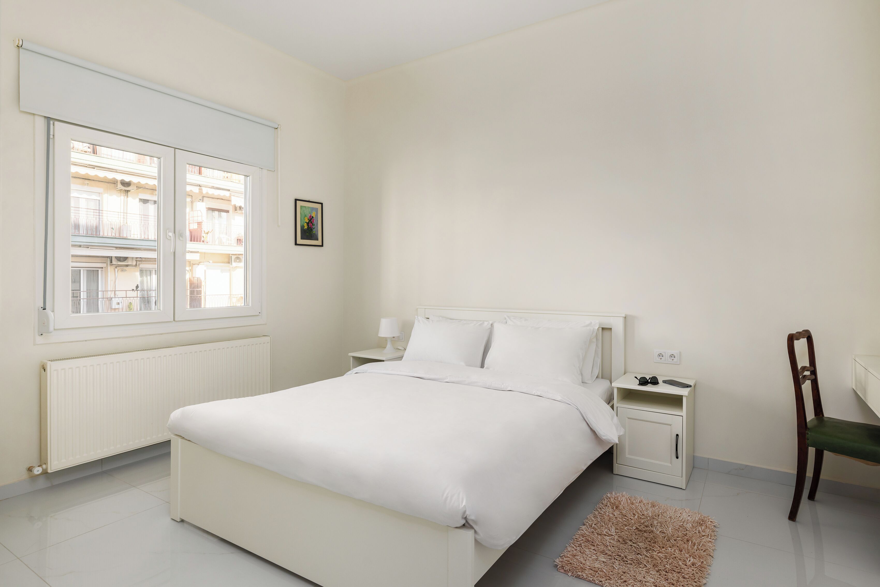 Una camera da letto con un letto, un comodino, una sedia, un termosifone e una finestra vista edifici.