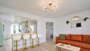 Une cuisine moderne avec des meubles blancs, un îlot central et des tabourets de bar. On y trouve également un coin repas avec une table en bois et un canapé orange.