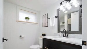 Ein modernes Badezimmer mit einer weißen Toilette, dunklen Holzschränken, einem großen, beleuchteten Spiegel und einem kleinen Fenster mit Pflanze.