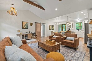Ein geräumiges Wohnzimmer mit einer Couch, Sesseln und einem Couchtisch. Im Hintergrund befindet sich ein Küchenbereich mit Schränken und einem Kühlschrank.